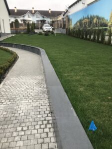 Long paved path with tightly packed Black Sea Blue Granite cobblestones and large curbstone wall adjacent to grass, surrounded by residential homes.