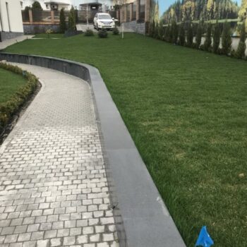 Long paved path with tightly packed Black Sea Blue Granite cobblestones and large curbstone wall adjacent to grass, surrounded by residential homes.