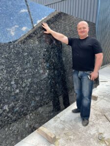 Standing Beside a Very Large Black Sea Blue Granite Slab, Showcasing its Scale and Surface Finish.