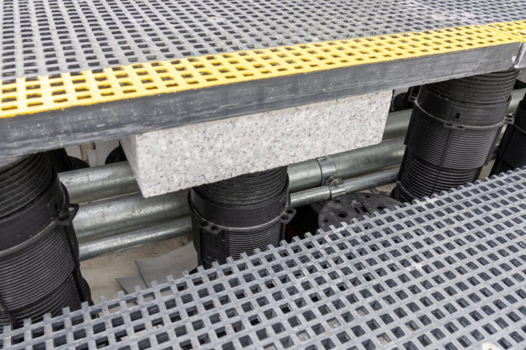 Close-up of grating panel edge with ballast and conduit visible beneath pedestal system