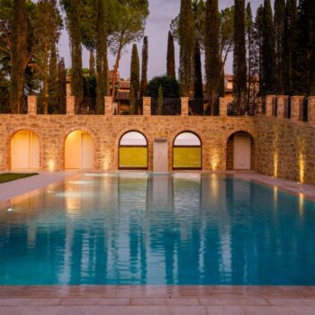 Nighttime view of a luxury pool surrounded by Rosca Gold porcelain pavers and uplighted stone architecture.