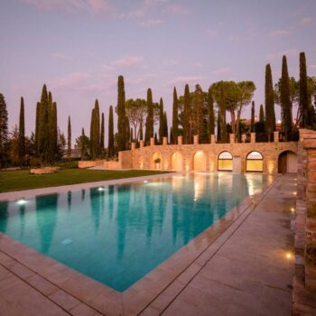 Wide nighttime view of a luxury pool featuring Rosca Gold porcelain pavers, accent lighting, and illuminated arched stone walls.